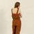 A light-skinned woman with long brown hair stands facing forward, wearing a linen menique jumpsuit with a tied belt at the waist and olive-green slip-on shoes. She has a neutral expression and her hands are resting on her hips.