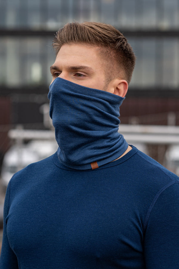 A man wearing the menique unisex denim neck gaiter. He is outside, wearing a matching color long sleeve merino top.